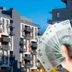 Dłoń trzyma banknoty 100 zł na tle nowoczesnych bloków mieszkalnych. Zastanawiasz się, ile kosztuje wynajem mieszkania w Polsce?