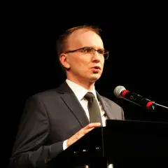 Mężczyzna w garniturze i okularach przemawia przy mikrofonie na czarnym tle.