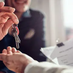 Przekazanie kluczy do nowego mieszkania. Uśmiechnięty mężczyzna wręcza pęk kluczy, a obok leży dokumentacja dotycząca tego, co ile płaci się czynsz.