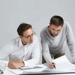 Dwóch mężczyzn analizuje plany, dyskutując, jak obliczyć kubaturę pomieszczenia. Jeden wskazuje na rysunek.