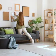 Kobieta wiesza ramki na ścianie, przygotowując mieszkanie do wynajmu. Potrzebne do wynajmu mieszkania: sofa, dekoracje, meble.