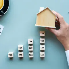 Dłoń układa drewniany domek na kostkach z napisem "TAX", symbolizując podatek od nieruchomości w Warszawie. Obok kalkulator i wykres.