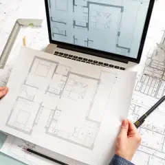 Ręce trzymające plan mieszkania, obok laptop z projektem, makieta domu i dokumentacja techniczna.
