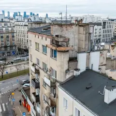 Widok na zaniedbany budynek mieszkalny z balkonami i panoramę miasta w tle.