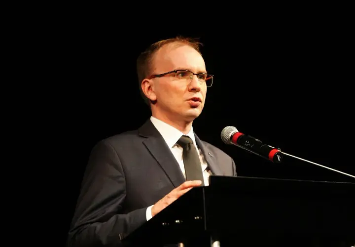 Mężczyzna w garniturze i okularach przemawia przy mikrofonie na czarnym tle.