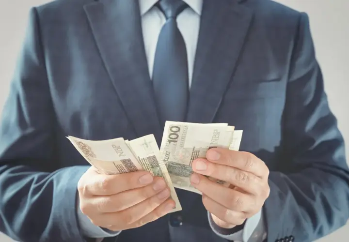 Mężczyzna w garniturze liczy banknoty 100 zł. Zastanawiasz się, ile zarabia przewodniczący rady gminy?