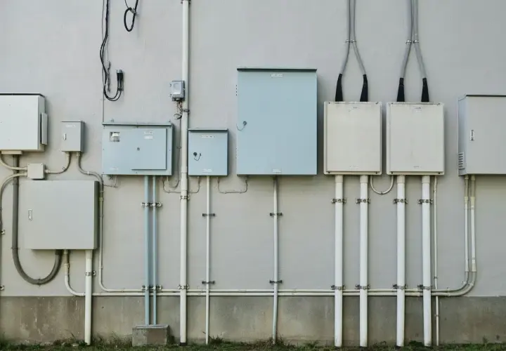 Szereg skrzynek elektrycznych i rur na ścianie budynku, przygotowanie do podłączenia prądu do działki bez domu.