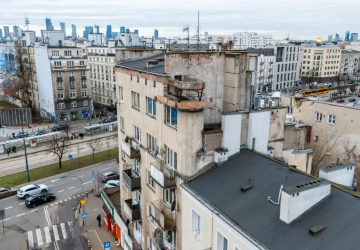 Widok na zaniedbany budynek mieszkalny z balkonami i panoramę miasta w tle.