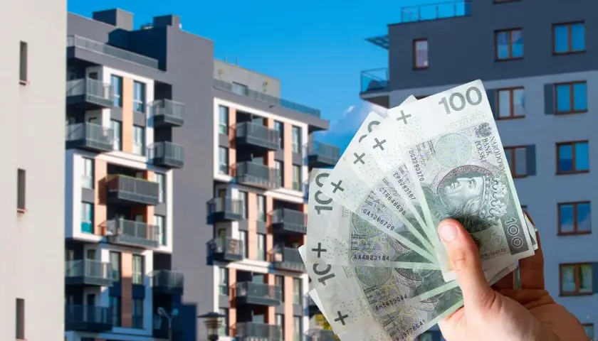 Dłoń trzyma banknoty 100 zł na tle nowoczesnych bloków mieszkalnych. Zastanawiasz się, ile kosztuje wynajem mieszkania w Polsce?