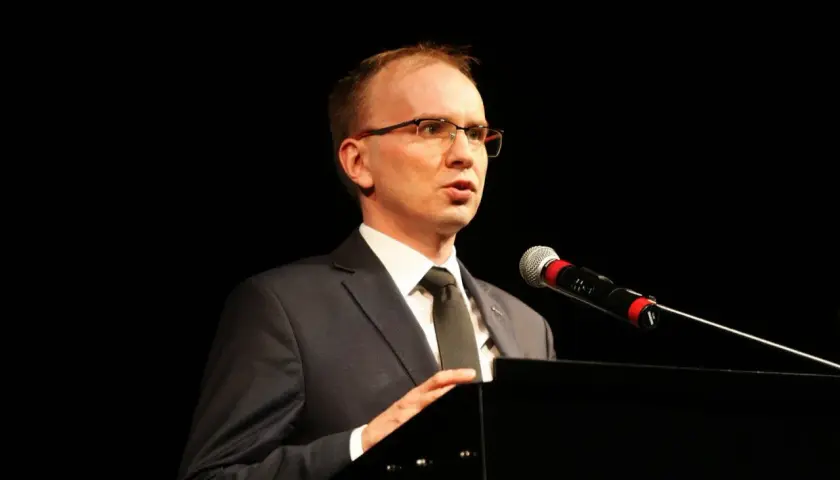 Mężczyzna w garniturze i okularach przemawia przy mikrofonie na czarnym tle.