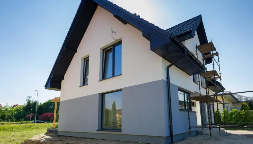Nowy dom z dwukolorową elewacją, w trakcie prac wykończeniowych. Widać rusztowania i fragment ogrodu.