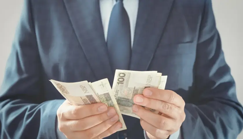 Mężczyzna w garniturze liczy banknoty 100 zł. Zastanawiasz się, ile zarabia przewodniczący rady gminy?