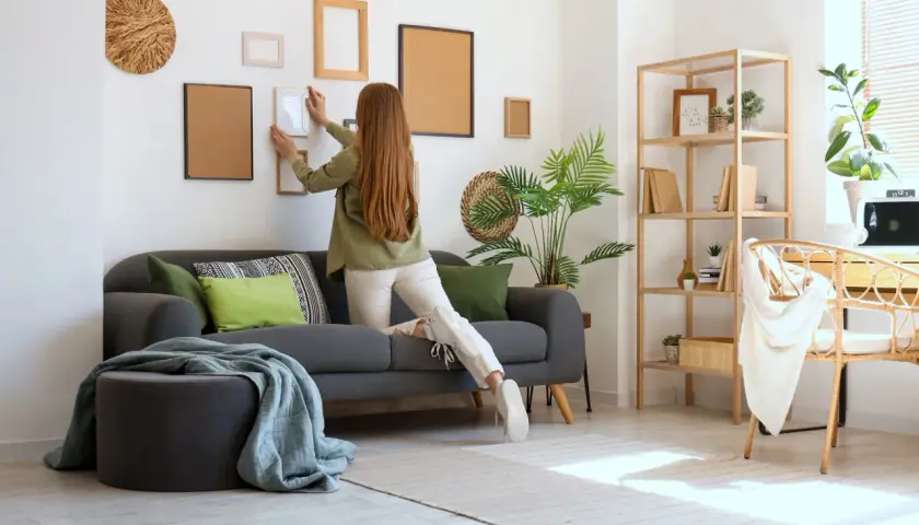Kobieta wiesza ramki na ścianie, przygotowując mieszkanie do wynajmu. Potrzebne do wynajmu mieszkania: sofa, dekoracje, meble.