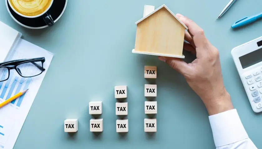 Dłoń układa drewniany domek na kostkach z napisem "TAX", symbolizując podatek od nieruchomości w Warszawie. Obok kalkulator i wykres.