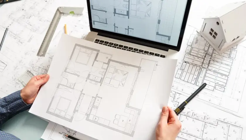 Ręce trzymające plan mieszkania, obok laptop z projektem, makieta domu i dokumentacja techniczna.