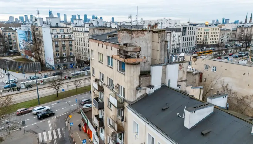 Widok na zaniedbany budynek mieszkalny z balkonami i panoramę miasta w tle.