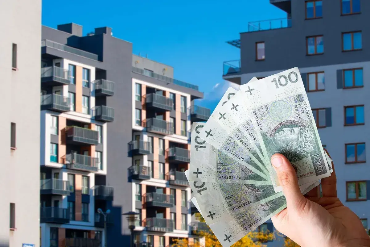 Dłoń trzyma banknoty 100 zł na tle nowoczesnych bloków mieszkalnych. Zastanawiasz się, ile kosztuje wynajem mieszkania w Polsce?