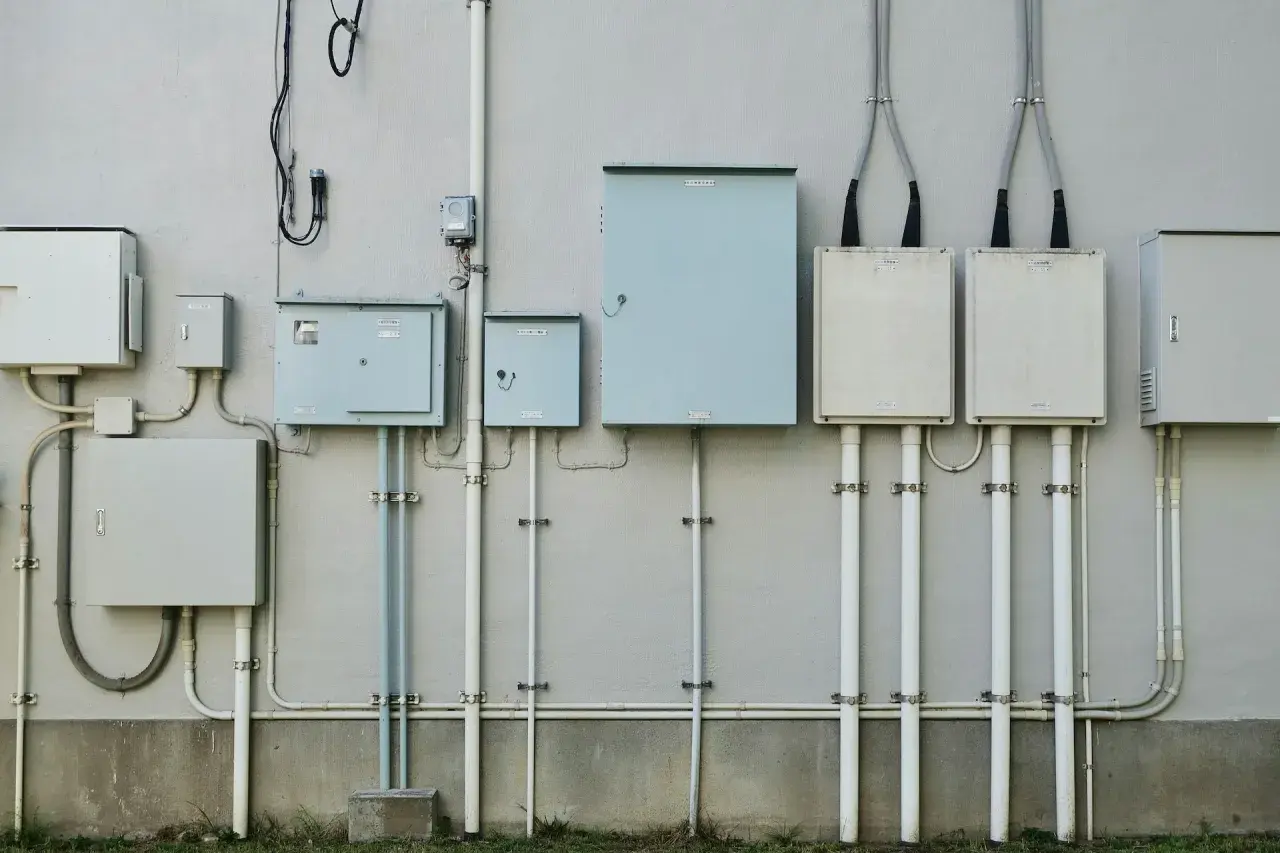 Szereg skrzynek elektrycznych i rur na ścianie budynku, przygotowanie do podłączenia prądu do działki bez domu.