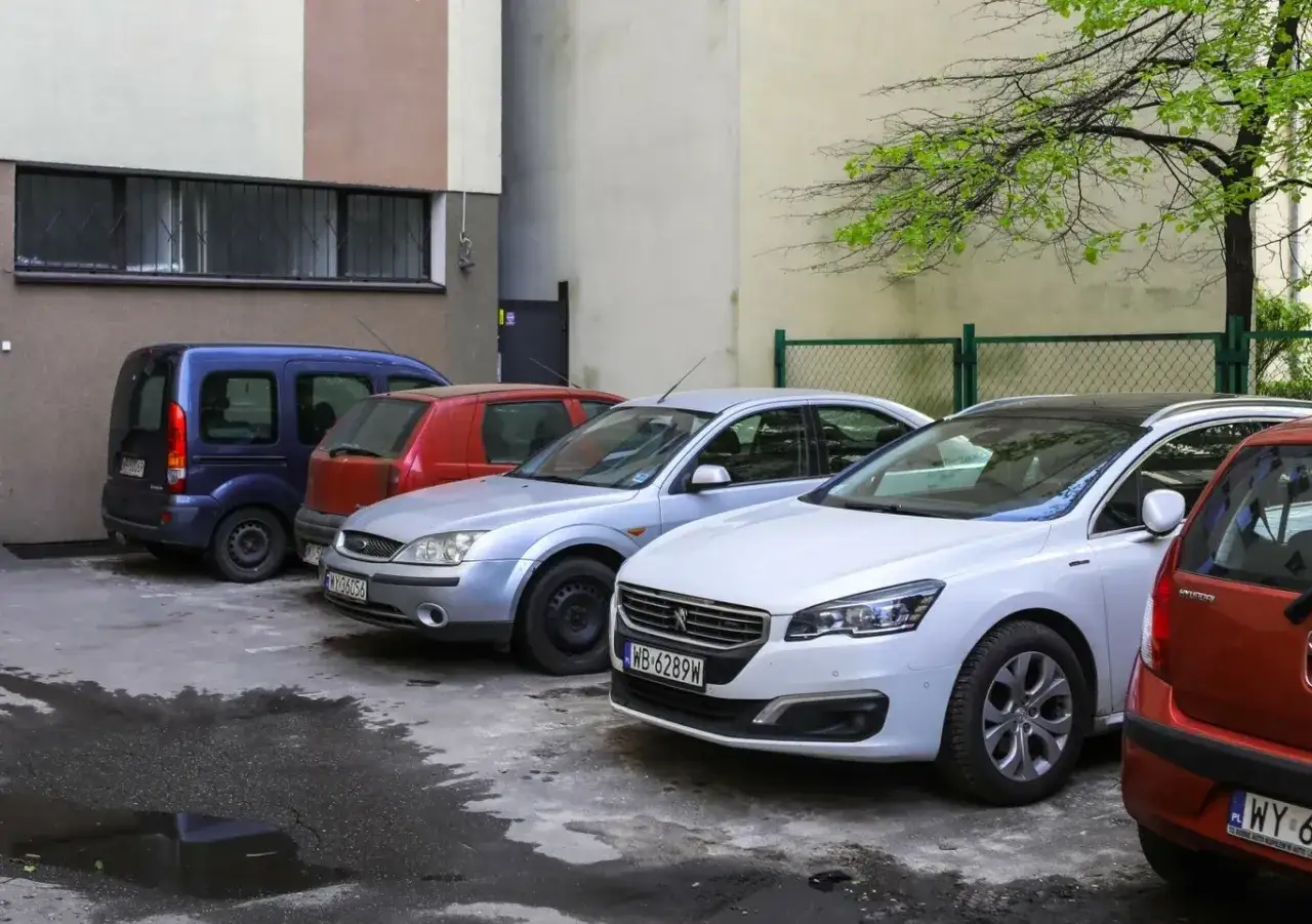 Samochody zaparkowane tyłem do bloku. Czy można parkować tyłem do bloku? Widać niebieskiego vana, czerwone auto i dwa srebrne samochody.