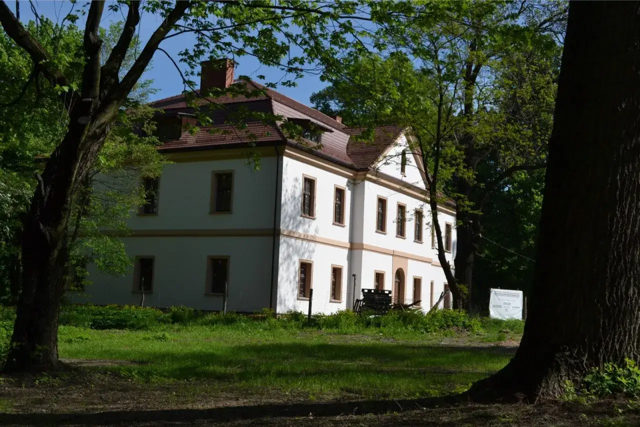 Biały dwór otoczony zielenią, kiedy dom jest zabytkiem. Wiosna w pełni, drzewa szumią wokół.