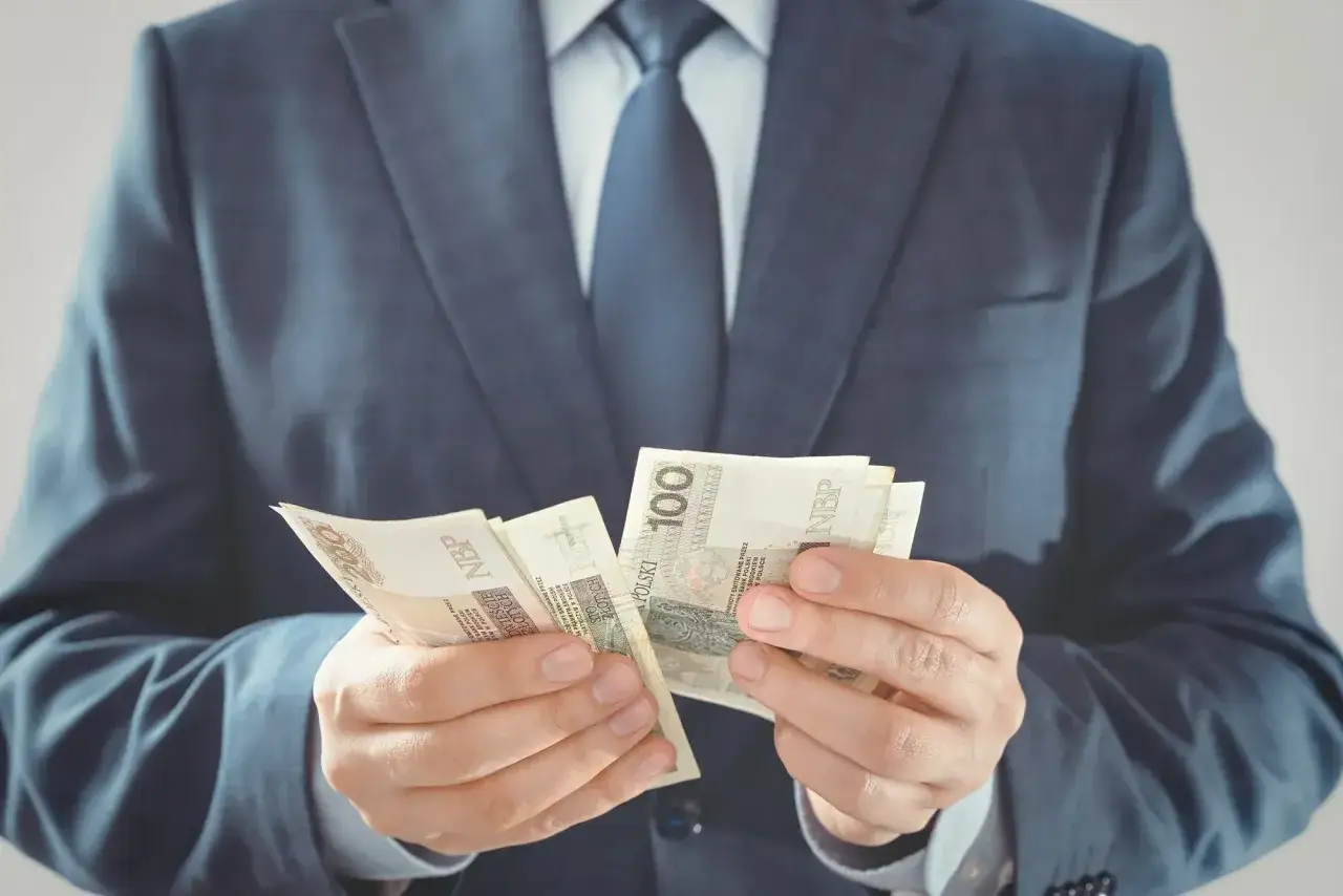 Mężczyzna w garniturze liczy banknoty 100 zł. Czy to jego pensja, ile zarabia przewodniczący rady gminy?