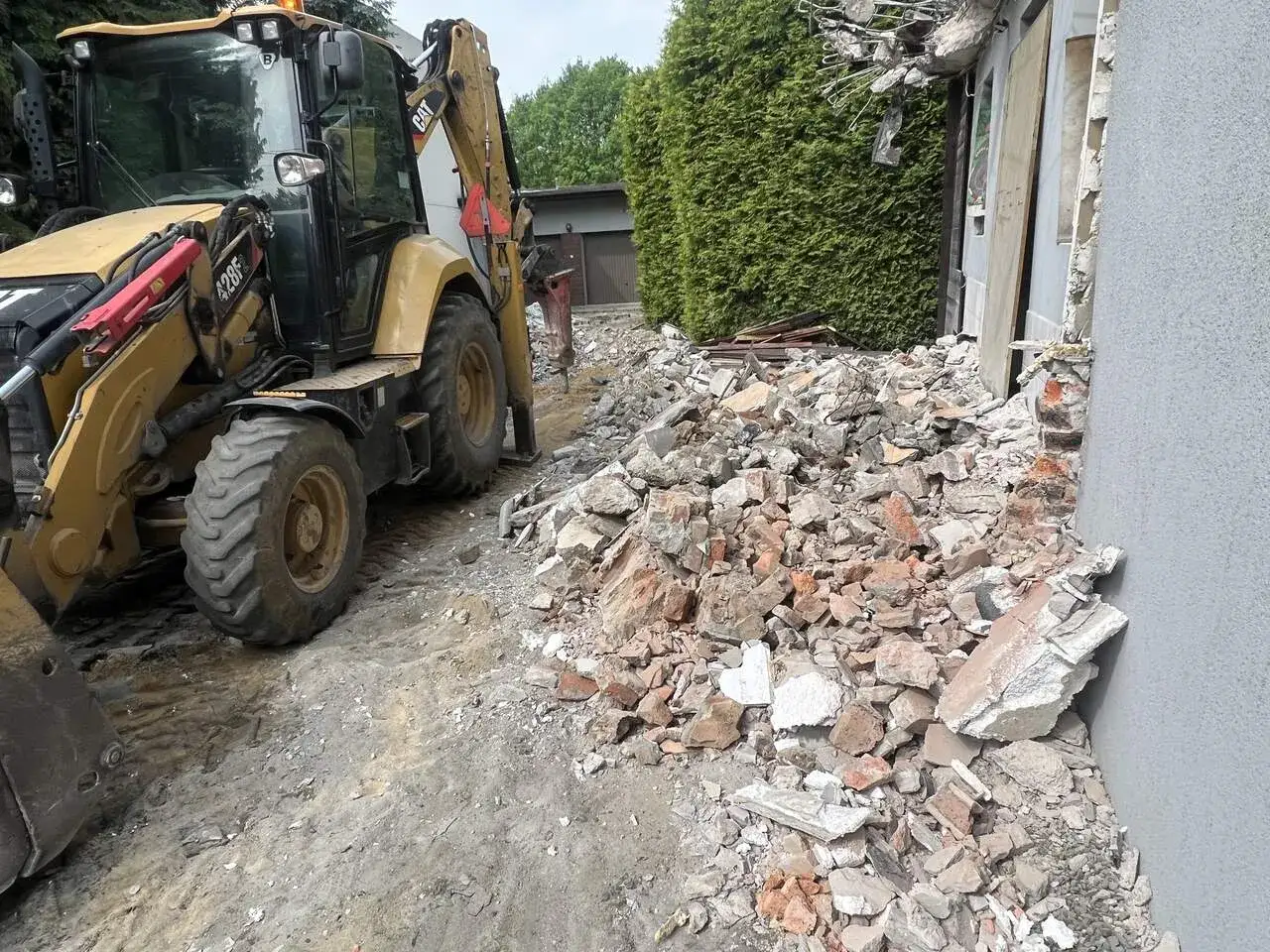 Żółta koparko-ładowarka pracuje przy rozbiórce starego domu. Gruz i cegły leżą na ziemi. Zastanawiasz się, ile kosztuje wyburzenie starego domu?