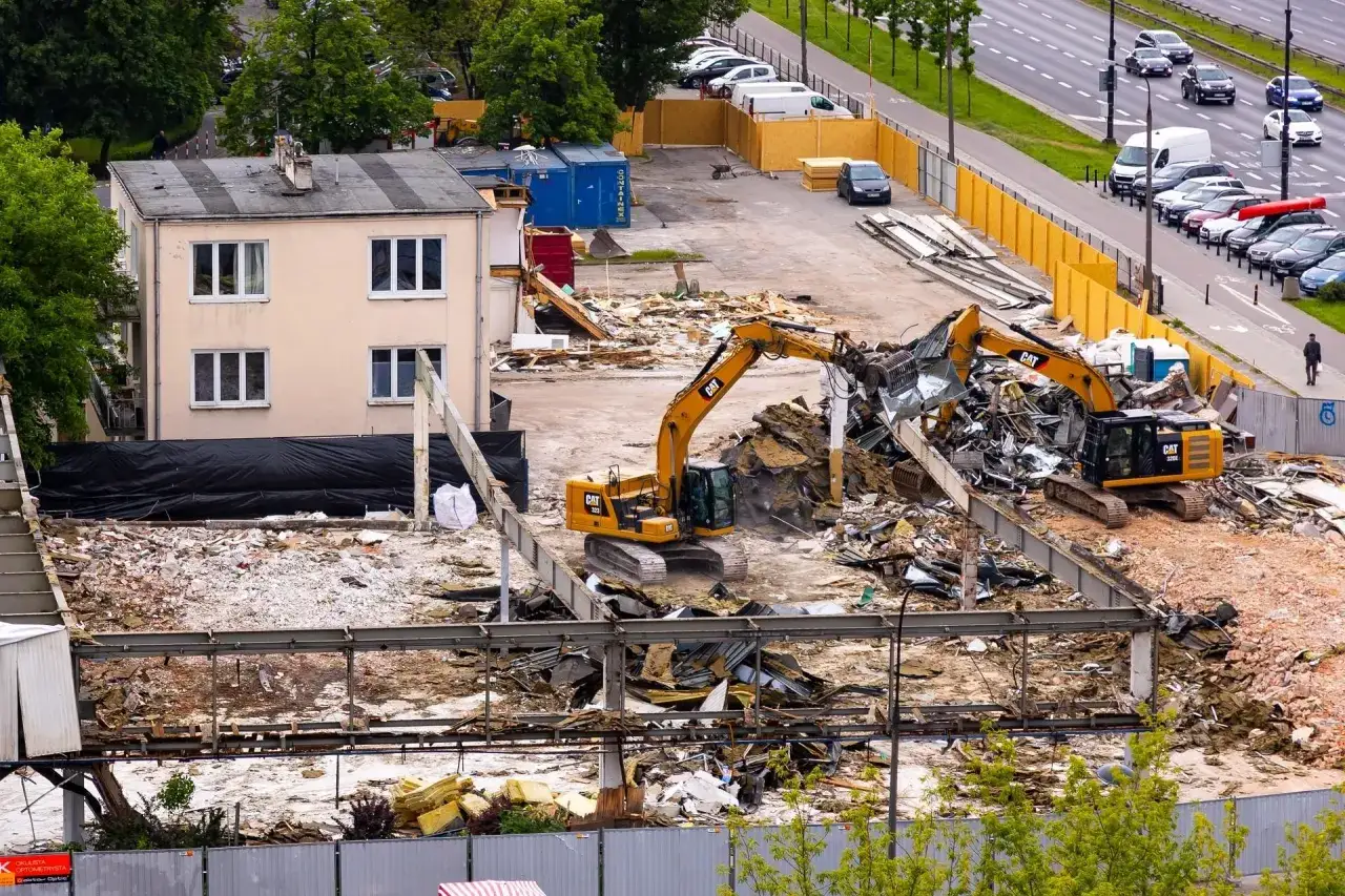 Dwie koparki pracują na placu budowy, gdzie trwa rozbiórka budynku. W tle widać ruchliwą ulicę i zaparkowane samochody. Warto wiedzieć, gdzie zgłosić rozbiórkę budynku, aby wszystko odbyło się zgodnie z prawem.