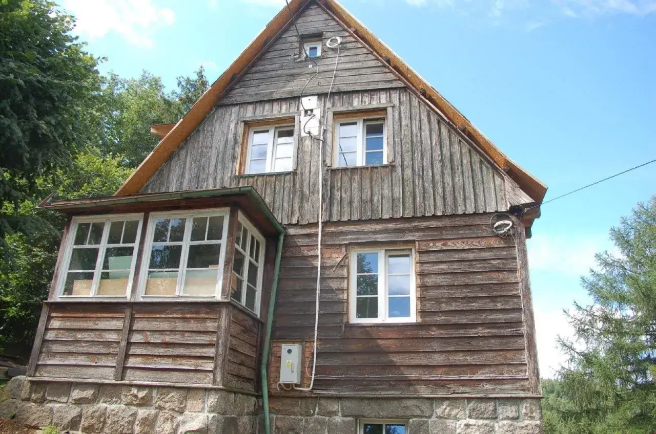 Drewniany dom z kamienną podmurówką i werandą, kiedy dom jest zabytkiem, otoczony zielenią.