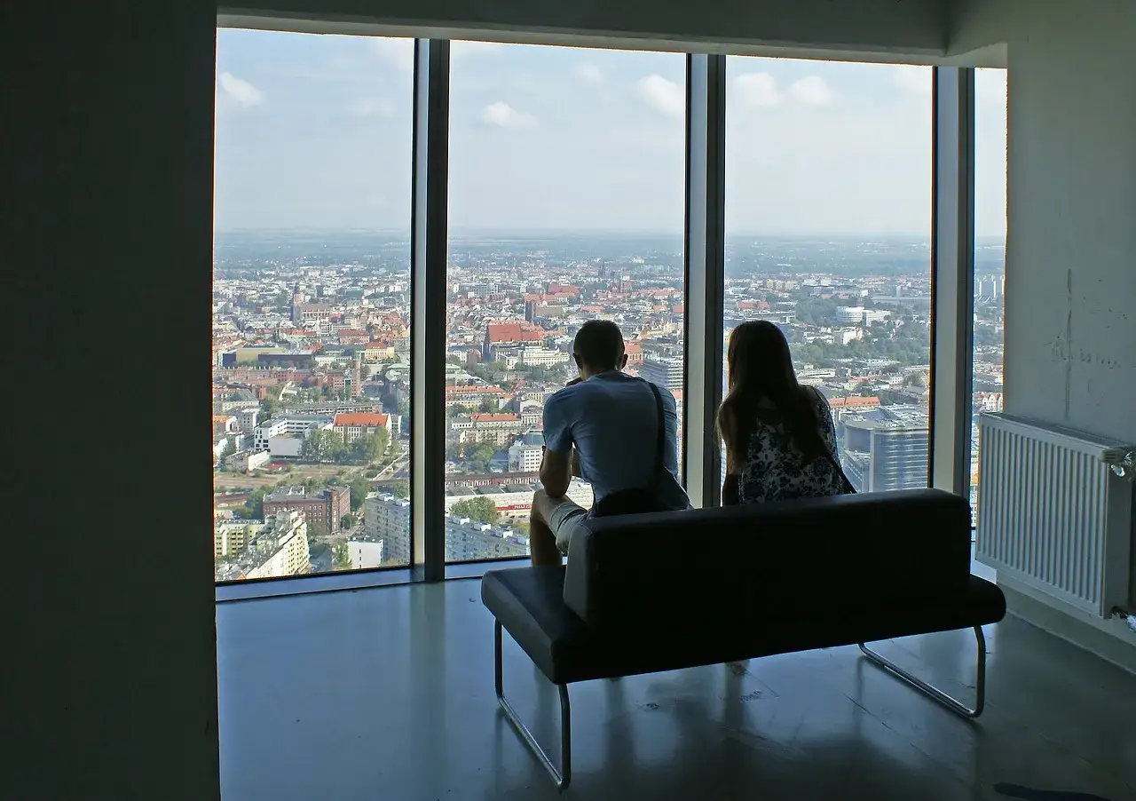 Para siedzi na kanapie, podziwiając panoramę miasta z okna. Zastanawiają się, ile Sky Tower ma pięter, by móc zobaczyć tak rozległy widok.